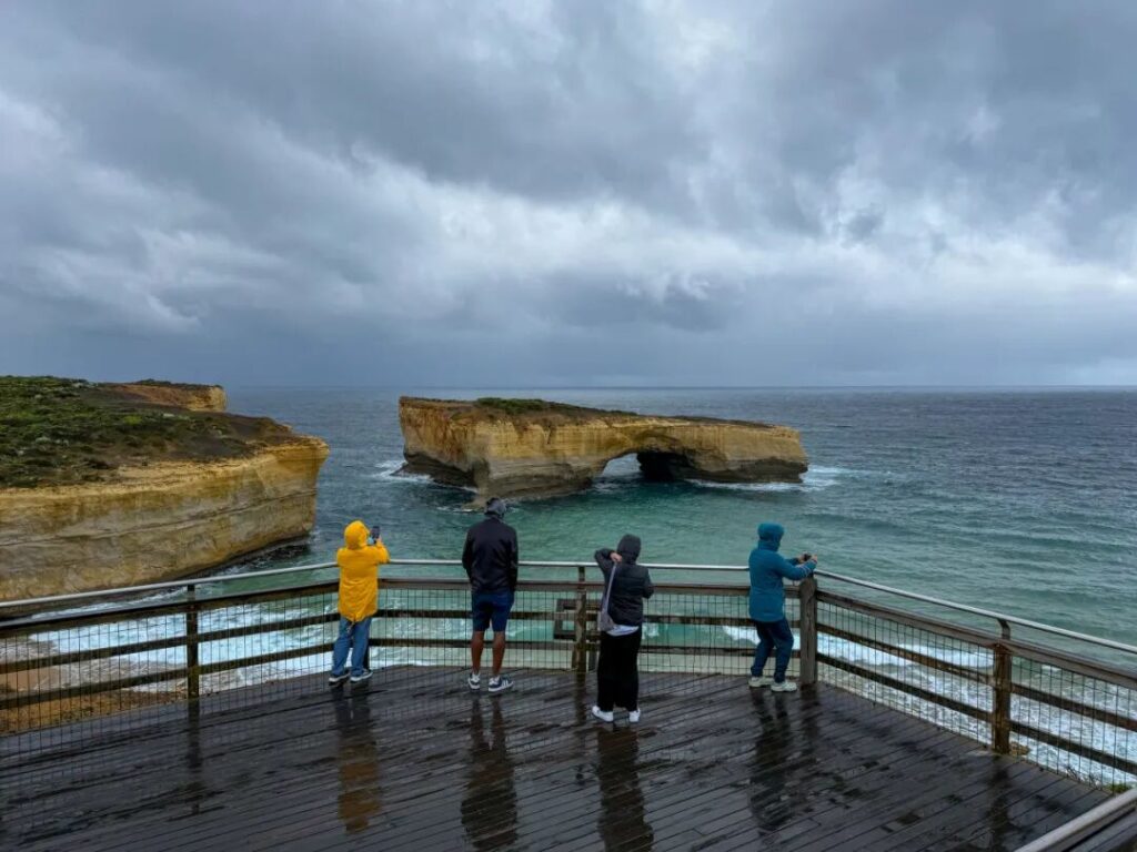 游客们在大洋路伦敦桥观景台上欣赏海岸风光，雨天穿着雨衣等待最佳光线